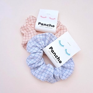 Könnte beinhalten: Zwei Haargummis, einer rosa und einer blau, beide mit einem Gittermuster. Jedes Haargummi hat ein weißes Etikett mit dem Wort "Pancho" und "LOVELY HANDMADE" unter einer Wimpernillustration.