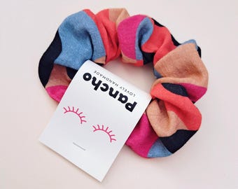 Könnte beinhalten: Ein Haargummi aus Stoff mit einem mehrfarbigen geometrischen Muster in Blau, Pink, Schwarz und Beige. Daran befestigt ist ein weißes rechteckiges Etikett mit dem Text "pancho LOVELY HANDMADE" und einer Grafik von zwei Wimpernpaaren.