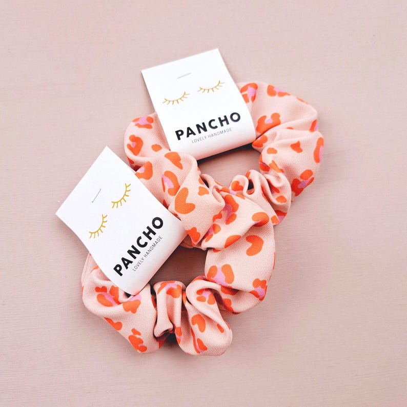 K&ouml;nnte beinhalten: Zwei Haargummis im Leopardenmuster in Rosa und Orange mit einem wei&szlig;en Etikett, auf dem "PANCHO LOVELY HANDMADE" steht.