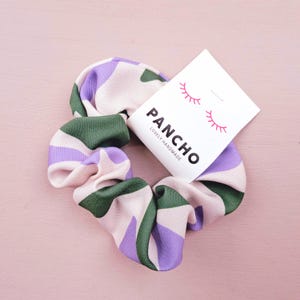 Könnte beinhalten: Ein pinkfarbenes, grünes und violettes Scrunchie mit einem weißen Etikett, auf dem "PANCHO LOVELY HANDMADE" steht und zwei rosa Wimpern.