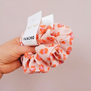 K&ouml;nnte beinhalten: Ein pinkfarbener und orangefarbener Scrunchie mit Leopardenmuster und einem wei&szlig;en Etikett mit der Aufschrift "PANCHO LOVELY HANDMADE".