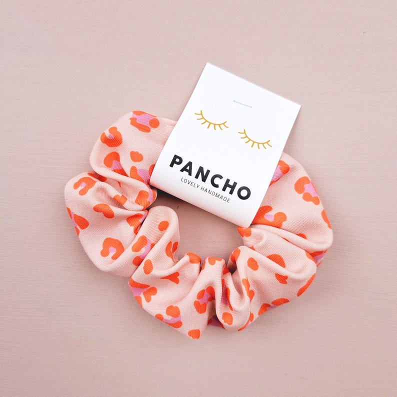 K&ouml;nnte beinhalten: Ein pinkfarbener und orangefarbener Scrunchie mit Leopardenmuster und einem wei&szlig;en Etikett mit der Aufschrift "PANCHO LOVELY HANDMADE" und zwei goldenen Wimpern.