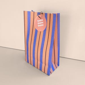 Könnte beinhalten: Eine Geschenktüte aus Papier mit vertikalen Streifen in Blau, Pfirsich und Dunkelbraun. Ein runder, pfirsichfarbener Aufkleber ist oben in der Mitte mit einer herzförmigen Metallklammer befestigt. Der Aufkleber trägt den Text happy happy happy in Weiß.