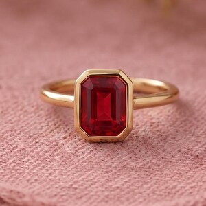 Puede incluir: Un anillo de oro con una piedra preciosa roja de corte esmeralda rectangular engastada en un bisel liso. La banda es fina y pulida, mostrando un acabado metálico limpio que complementa el color rojo vibrante y profundo de la piedra central.