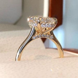 Puede incluir: Un anillo de compromiso de oro con una gran piedra central de diamante de talla ovalada. El anillo presenta un halo de diamantes m&aacute;s peque&ntilde;os alrededor de la piedra central y a lo largo de la banda. El anillo est&aacute; engastado en oro.