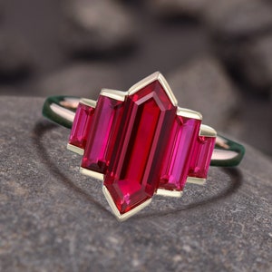 May include: A gold-toned ring featuring a geometric design with a large, rectangular, deep red gemstone at the center, flanked by two smaller, matching stones. The ring is set on a gray stone surface, with the text "foreverjewellstudio" at the top.