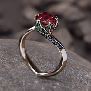 Könnte beinhalten: Ein rosengestaltiger Ring mit einem roten Edelstein, der von grünen Edelsteinen umgeben ist. Die Ringschiene ist aus Gold und hat schwarze Edelsteine.