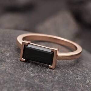 May include: A rose gold ring with a rectangular black gemstone. The ring is set on a gray stone surface. The text "Foreverjewellstudio" is at the top of the image.