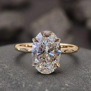 Puede incluir: Un anillo de compromiso dorado con un diamante de talla ovalada. El anillo est&aacute; sobre una piedra gris, con el texto "Foreverjewellstudio" en la parte superior. El diamante es grande y claro, reflejando la luz.