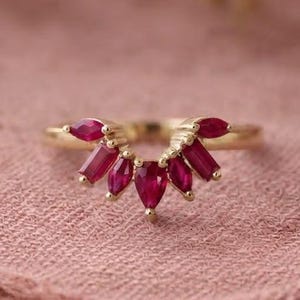 May include: A gold ring featuring a curved band set with five vibrant red gemstones. The stones are arranged in a fan-like pattern, consisting of two marquise-cut stones on the outer edges, two baguette-cut stones, and a central pear-cut gemstone.