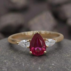 Peut inclure: Une bague en or avec une pierre centrale rouge rubis en forme de poire et deux pierres latérales blanches en forme de poire. La bague est posée sur une pierre grise. Le texte "Foreverjewellstudio" est en haut.
