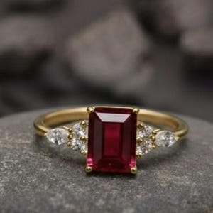 Może przedstawiać: Złoty pierścionek z prostokątnym, głęboko czerwonym kamieniem szlachetnym jako centralnym elementem, otoczonym mniejszymi, białymi kamieniami szlachetnymi w kształcie gruszki. Pierścionek leży na szarej kamiennej powierzchni, z tekstem "foreverjewellstudio" u góry.