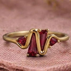 Puede incluir: Un anillo de oro con una banda ondulada y sinuosa engastada con cuatro piedras preciosas facetadas de color rojo intenso. Las piedras incluyen dos formas trapezoidales alargadas en el centro y dos piedras triangulares más pequeñas en los bordes.