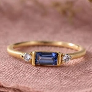 Op de afbeelding: Een gouden ring met een centrale rechthoekige blauwe saffier in baguette-slijpvorm, geflankeerd door twee kleine ronde heldere stenen. De ringband is dun en gepolijst, met een strak ontwerp en een veilige zetting voor de drie edelstenen.