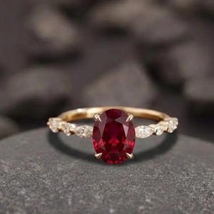May include: A gold ring with a large oval ruby center stone. The band features small, clear accent stones. The ring is set on a gray stone surface. The text "Foreverjewellstudio" is at the top.
