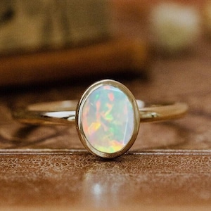 Könnte beinhalten: Ein goldener Ring mit einem weißen Opal-Edelstein. Der Ring liegt auf einem braunen ledergebundenen Buch.