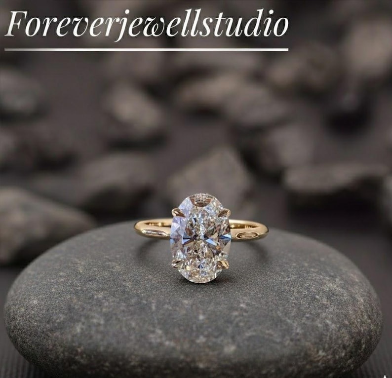 Puede incluir: Un anillo de compromiso dorado con un diamante de talla ovalada. El anillo est&aacute; sobre una piedra gris, con el texto "Foreverjewellstudio" en la parte superior. El diamante es grande y claro, reflejando la luz.