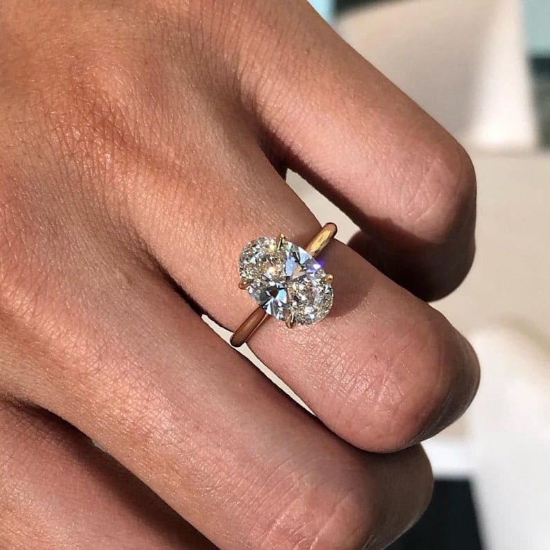 Puede incluir: Un anillo de oro con un gran diamante de talla ovalada. El diamante brilla con numerosas facetas, reflejando la luz. La banda del anillo es de color oro c&aacute;lido, y el engaste sujeta la gema de forma segura. El anillo se muestra en un dedo.