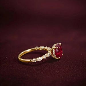 K&ouml;nnte beinhalten: Ein goldener Ring mit einem gro&szlig;en, oval geschliffenen Rubin als Mittelpunkt. Der Ring hat ein zartes Band mit kleinen, funkelnden Akzentsteinen. Der Rubin hat eine tiefrote Farbe, und der Ring ist vor einem dunkelroten Hintergrund platziert.