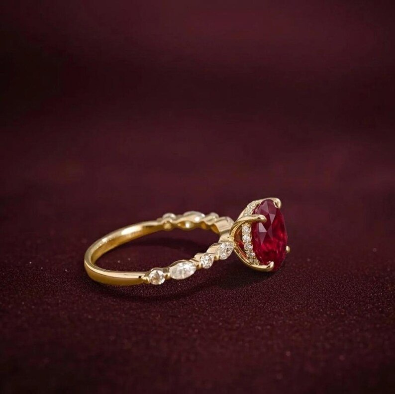 K&ouml;nnte beinhalten: Ein goldener Ring mit einem gro&szlig;en, oval geschliffenen Rubin als Mittelpunkt. Der Ring hat ein zartes Band mit kleinen, funkelnden Akzentsteinen. Der Rubin hat eine tiefrote Farbe, und der Ring ist vor einem dunkelroten Hintergrund platziert.