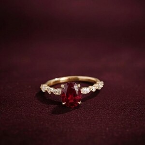 K&ouml;nnte beinhalten: Ein goldener Ring mit einem gro&szlig;en, oval geschliffenen, tiefroten Edelstein als Mittelpunkt. Das Band ist mit kleineren, klaren Edelsteinen verziert. Der Ring ist vor einem dunklen burgunderfarbenen Hintergrund platziert, der das Funkeln und Design des Schmucks hervorhebt.