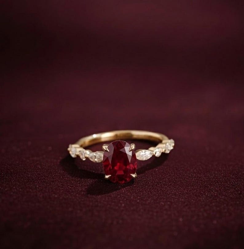 K&ouml;nnte beinhalten: Ein goldener Ring mit einem gro&szlig;en, oval geschliffenen, tiefroten Edelstein als Mittelpunkt. Das Band ist mit kleineren, klaren Edelsteinen verziert. Der Ring ist vor einem dunklen burgunderfarbenen Hintergrund platziert, der das Funkeln und Design des Schmucks hervorhebt.