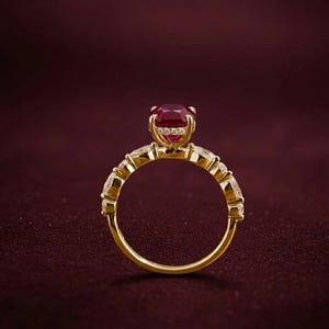 K&ouml;nnte beinhalten: Ein goldener Ring mit einem gro&szlig;en, ovalen, tiefroten Edelstein als Mittelpunkt. Der Ring ist mit kleineren, klaren Edelsteinen besetzt und hat eine polierte Oberfl&auml;che. Der Ring ist vor einem dunkelroten Hintergrund platziert.