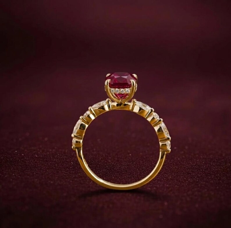 K&ouml;nnte beinhalten: Ein goldener Ring mit einem gro&szlig;en, ovalen, tiefroten Edelstein als Mittelpunkt. Der Ring ist mit kleineren, klaren Edelsteinen besetzt und hat eine polierte Oberfl&auml;che. Der Ring ist vor einem dunkelroten Hintergrund platziert.