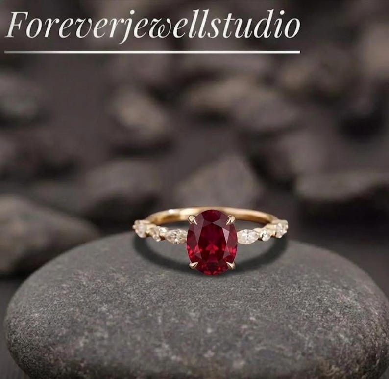 K&ouml;nnte beinhalten: Ein goldener Ring mit einem gro&szlig;en ovalen Rubin-Mittelstein. Das Band hat kleine, klare Akzentsteine. Der Ring liegt auf einer grauen Steinoberfl&auml;che. Der Text "Foreverjewellstudio" steht oben.