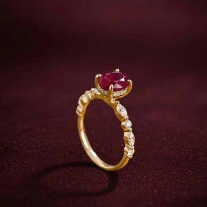 K&ouml;nnte beinhalten: Ein goldener Ring mit einem gro&szlig;en, oval geschliffenen Rubin als Mittelpunkt. Der Ring hat kleinere, klare Edelsteine und ein zartes Band. Der Rubin hat eine tiefrote Farbe, und der Ring ist vor einem dunkelburgunderfarbenen Hintergrund platziert.
