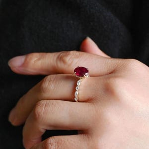 K&ouml;nnte beinhalten: Ein goldener Ring mit einem gro&szlig;en, ovalen Rubin. Das Band ist mit kleineren, klaren Edelsteinen verziert. Der Ring ist vor einem dunklen Hintergrund platziert, der das leuchtende Rot des Rubins hervorhebt. Ein Schmuckst&uuml;ck.