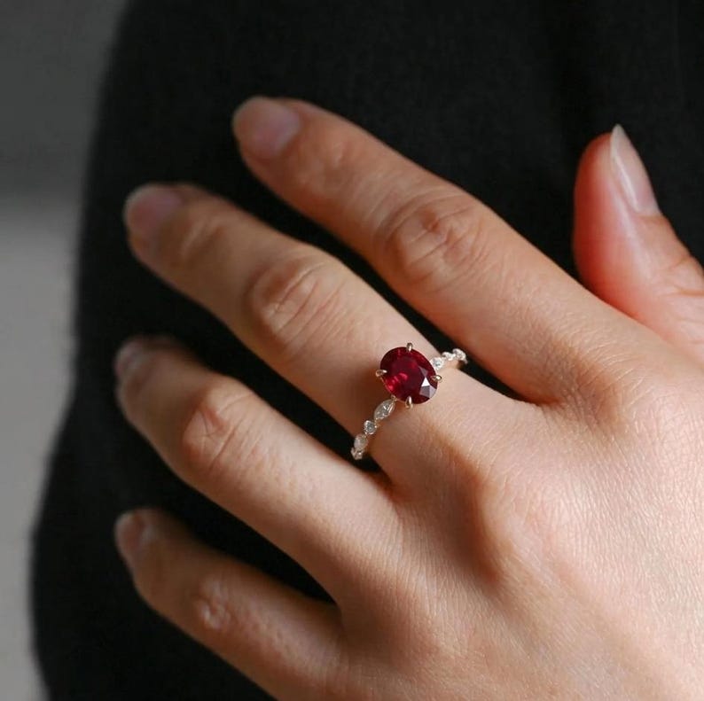 K&ouml;nnte beinhalten: Ein goldener Ring mit einem ovalen, tiefroten Edelstein als Mittelpunkt. Das Band ist mit kleinen, klaren Akzentsteinen verziert. Der Ring befindet sich an einer Hand, die sein Design und seine Farbe vor dunklem Hintergrund zeigt.