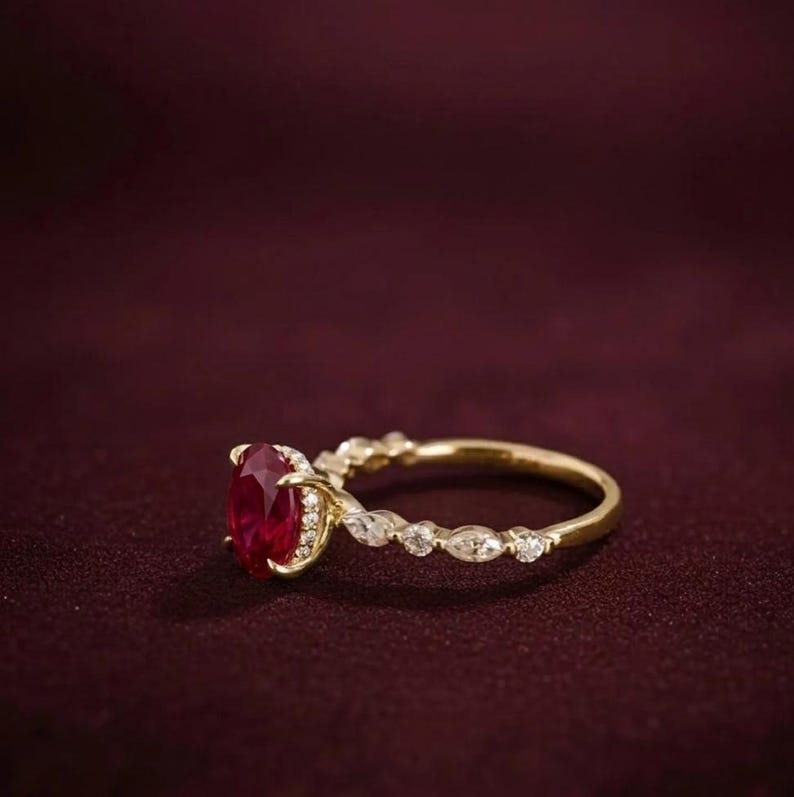 K&ouml;nnte beinhalten: Ein goldener Ring mit einem gro&szlig;en, oval geschliffenen Rubin als Mittelpunkt. Das Band weist kleine, klare Akzentsteine und ein filigranes Design auf. Der Ring ist vor einem dunkelroten Hintergrund platziert.
