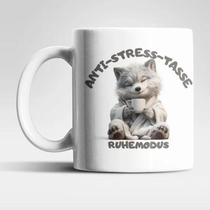 Könnte beinhalten: Ein weißer Keramikbecher mit einer Grafik eines flauschigen grauen Wolfs im Bademantel, der eine kleine weiße Tasse hält, mit dem Text "ANTI-STRESS-TASSE" darüber und "RUHEMODUS" unter dem Wolf in einer dunkelgrauen serifenlosen Schrift.