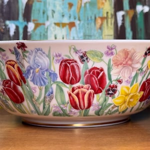 May include: A large, round, white ceramic bowl featuring a colorful botanical illustration of red tulips, blue irises, yellow daffodils, and small purple violets. The bowl has a smooth, glossy finish and a thin gold rim along the top and base edges.