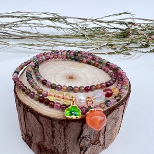 Puede incluir: Varios brazaletes de cuentas con una variedad de piedras preciosas coloridas, incluyendo piedras rosas, verdes y amarillas. Los brazaletes están adornados con dijes, incluyendo una flor verde y un colgante en forma de melocotón. Los brazaletes se exhiben sobre una rodaja de madera.