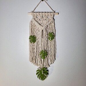 May include: A macrame wall hanging with a triangular shape and fringe. It features four green felt leaves with a gold glitter accent.