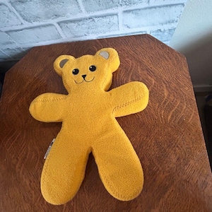 May include: A plush, yellow teddy bear-shaped pillow with a smiling face and small gray accents on the ears. The bear is lying on a brown wooden surface.