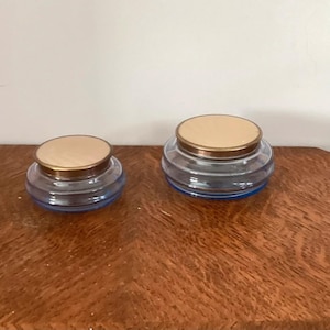 May include: Two round glass jars with blue-tinted bases and metallic gold-toned lids. The jars feature a stepped, tiered design on the glass body. One jar is smaller in diameter than the other, and both have a smooth, flat circular top surface.