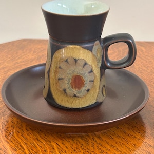 May include: A ceramic espresso cup and saucer set. The cup is dark brown with a light green interior and a handle. The cup is decorated with a floral pattern in shades of yellow and brown. The saucer is a matching dark brown colour.
