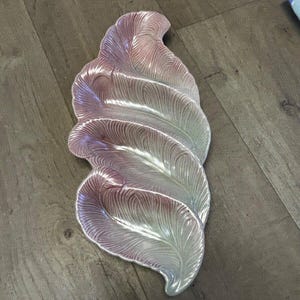 May include: An ornamental serving platter shaped like layered feathers. The dish has a glossy finish with a colour gradient, shifting from pale pink to silvery-white. The feather details are subtly textured, with a ridged pattern.