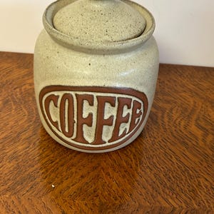 May include: A beige ceramic coffee canister with a lid. The word "COFFEE" is displayed in a brown oval on the front of the canister. The canister has a round shape and a small knob on the lid.