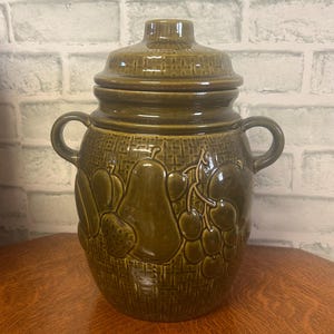May include: A green ceramic cookie jar with a lid and two handles. The jar is decorated with embossed fruit designs, including pears, cherries, and strawberries. The jar is sitting on a wooden surface.