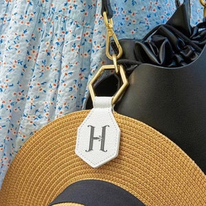 May include: A white leather bag charm with a gold hexagonal ring and the letter "H" in black script. The charm is attached to a black leather bag.