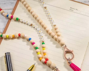 Puede incluir: Dos collares de cuentas de madera con letras doradas y borlas rosas. Los collares están sobre un cuaderno blanco con líneas y un bolígrafo negro y un bolígrafo plateado.