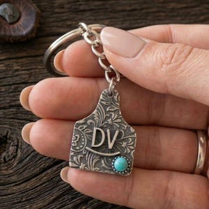 May include: A silver-colored keychain with a decorative tag featuring the initials "DV" and a small turquoise stone. The tag has an ornate, engraved pattern. The keychain is attached to a silver ring and held by a person.