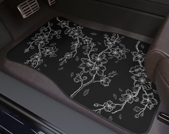May include: Black car floor mat with a white floral design. The design features delicate branches with white flowers and leaves.