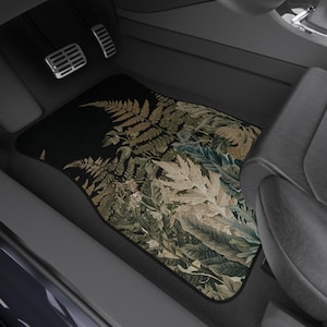 May include: A rectangular car floor mat featuring a detailed botanical print of various fern leaves and foliage in shades of dark green, muted gold, and beige against a solid black background, positioned in the footwell of a vehicle interior.