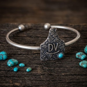 May include: A silver cuff bracelet featuring a central tag-shaped charm with an intricate floral pattern and the letters DV engraved on the front. A small, round turquoise stone is set into the lower right corner of the charm.
