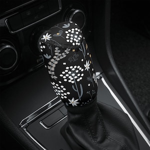 May include: Black gear shift knob cover with a white floral pattern. The pattern features small white flowers, leaves, and dots on a black background.